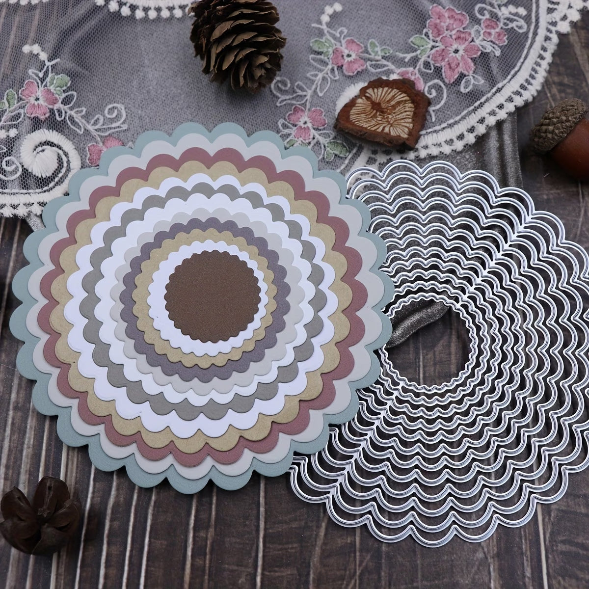 A collection of muted paper circles, cut with the Printable Heaven 12-Piece Scalloped Circle Cutting Die Set (D140), is artfully arranged next to nested black and white die-cut circles on a wooden surface—an inspiring setup for cardmaking or scrapbooking. Pinecones and floral embroidered lace in the background add a quaint, charming touch.