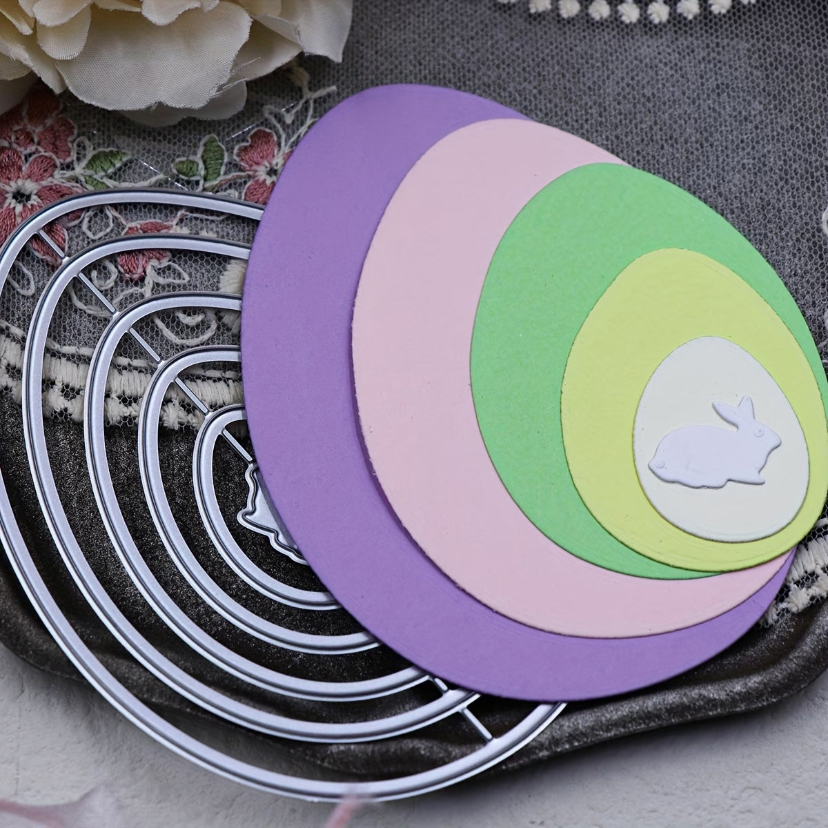 Six metal Nesting Eggs with Baby Rabbit Cutting Die (A097) by Printable Heaven are arranged alongside pastel paper ovals in purple, pink, green, and yellow, each size nesting into the next. The smallest oval showcases a white baby rabbit die-cut silhouette. Floral accents and lace appear in the background—an ideal scene for Easter paper crafting inspiration.