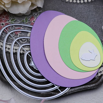 Six metal Nesting Eggs with Baby Rabbit Cutting Die (A097) by Printable Heaven are arranged alongside pastel paper ovals in purple, pink, green, and yellow, each size nesting into the next. The smallest oval showcases a white baby rabbit die-cut silhouette. Floral accents and lace appear in the background—an ideal scene for Easter paper crafting inspiration.