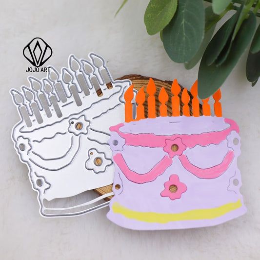 A Printable Heaven Birthday Cake Cutting Die (1 PC) (D049) is displayed alongside a vibrant paper cutout of a birthday cake with candles, pink icing, and decorative flowers on a wooden surface with green leaves nearby—ideal for card making or scrapbooking projects.