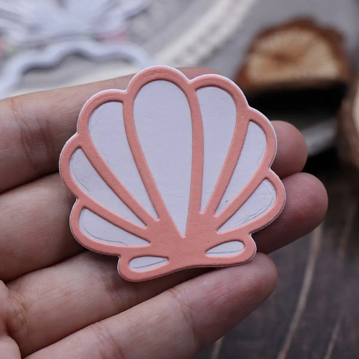 A hand holds a decorative, scallop-shaped cutout—crafted using the Printable Heaven Scallop Seashell Cutting Die (2 PCS) (T234)—featuring a pink outline and white interior. The blurred background displays additional craft materials, making it ideal for beach-themed crafts or card making projects.