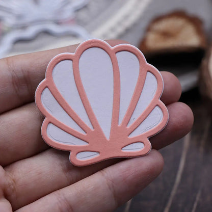 A hand holds a decorative, scallop-shaped cutout—crafted using the Printable Heaven Scallop Seashell Cutting Die (2 PCS) (T234)—featuring a pink outline and white interior. The blurred background displays additional craft materials, making it ideal for beach-themed crafts or card making projects.