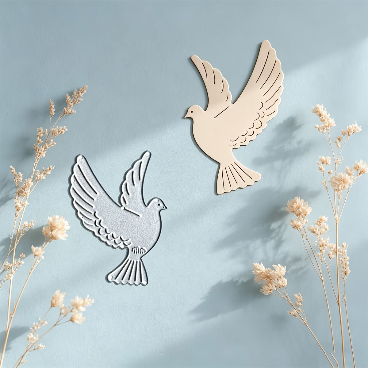 Adorn your creative projects with inspiration from this serene scene: a silver and a beige dove-shaped decoration mounted on a light blue wall, soft shadows cast by delicate dried flowers in the foreground. Perfect as inspiration for your next card or craft project using the Printable Heaven Flying Dove Metal Cutting Die (1 pc, A020).