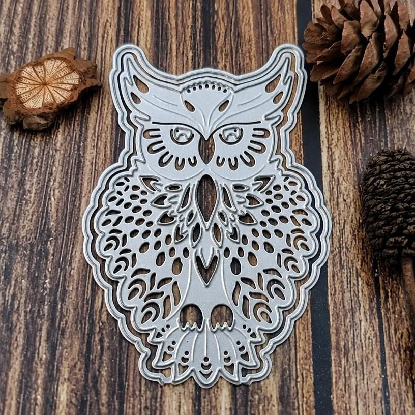 A Filigree Owl Cutting Die (1 PC) (A109) by Printable Heaven, featuring intricate metal cutout patterns, lies on a wooden surface beside pine cones and a small, round wood slice—ideal for card making or paper crafts.