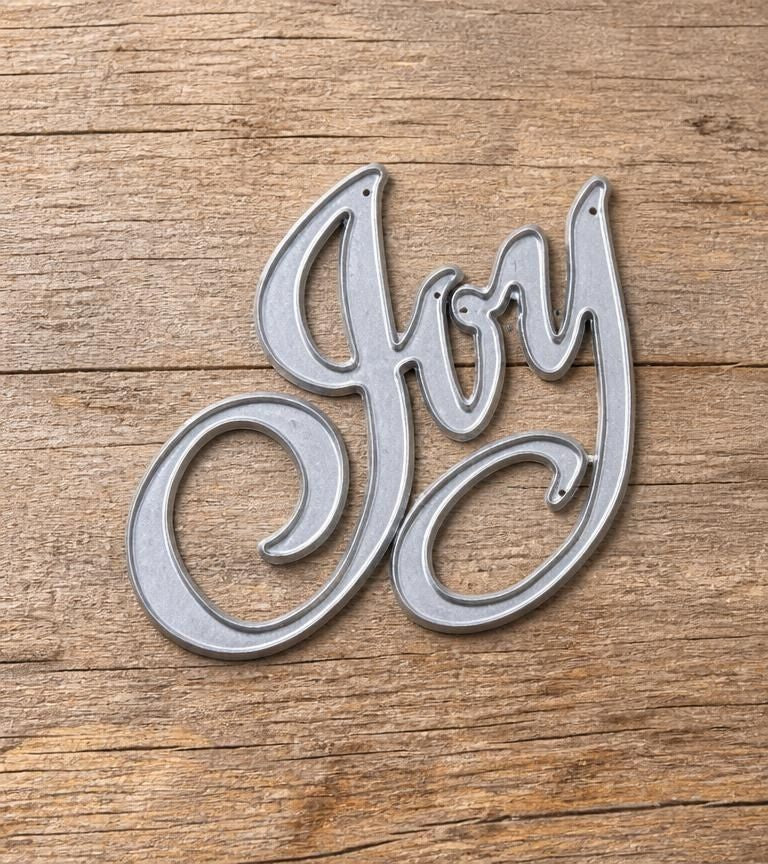 The Printable Heaven Joy Sentiment Cutting Die (1 PC) (W085) creates a silver metal script “Joy” that looks beautiful on a rustic wooden surface, making it ideal for adding a festive touch to handmade cards or paper crafting projects.