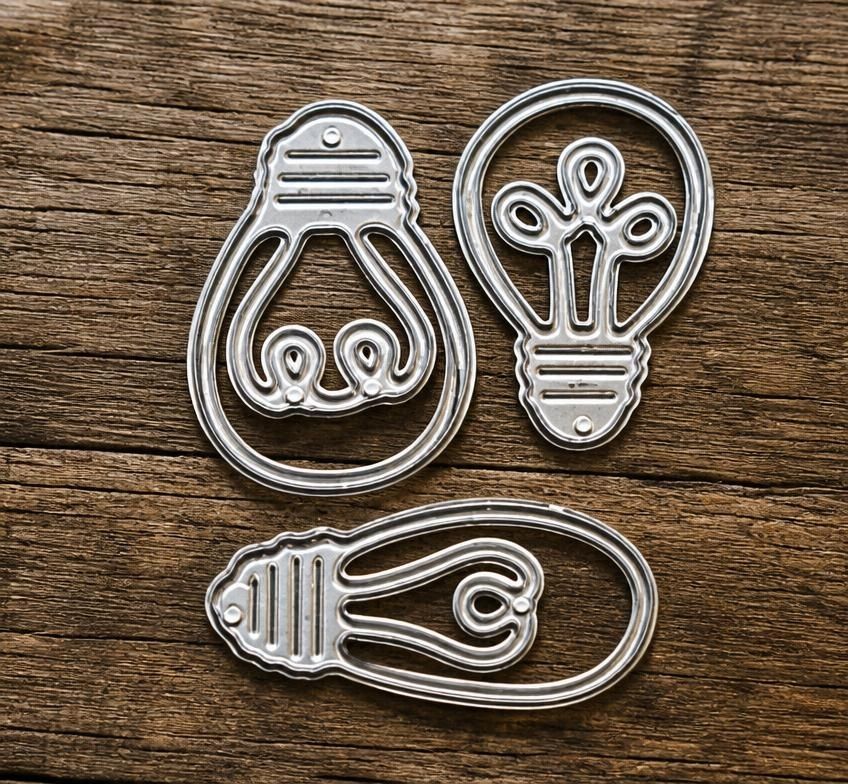 Three metal dies from the Printable Heaven Light Bulb & Energy-Themed Cutting Die Set (3 PCS) (D096) are arranged on a wooden surface in upright, tilted, and horizontal positions. Ideal for paper crafts and handmade cards, these versatile dies add a creative energy-inspired touch to any project.