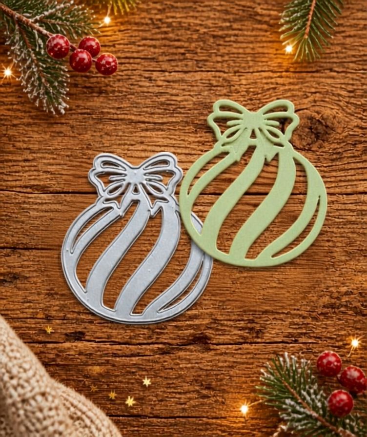 Two ornament-shaped cutouts—one in silver and one in green—rest on a wooden surface amidst pine branches, red berries, and festive lights, setting the scene for holiday crafting with the Printable Heaven Candy Cane Striped Christmas Bauble Cutting Die (1 PC) (SP005).