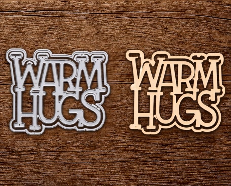 Two cutouts created with the Printable Heaven Warm Hugs Sentiment Cutting Die (2 PCS) (W071) rest on a wooden surface; the left cutout is silver-gray and the right is light brown, both featuring a playful outlined font style—ideal embellishments for handmade cards or scrapbooking projects to share heartfelt “Warm Hugs” sentiments.