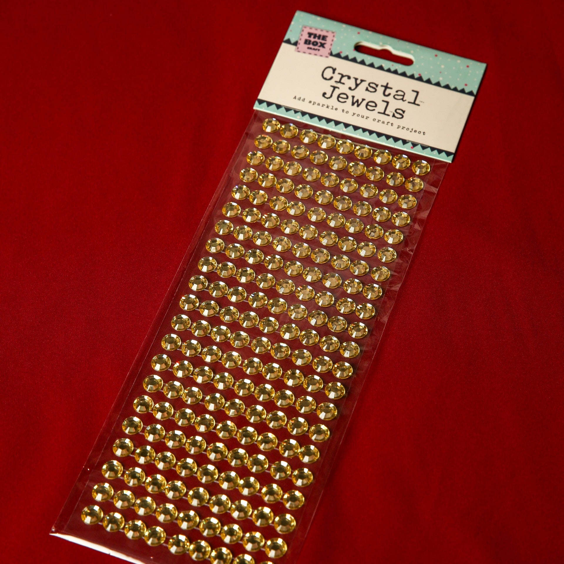 A pack of Printable Heaven 10mm Gold Crystal Adhesive Jewels is shown on a red surface. The sparkling gems, ideal for embellishing Christmas cards, are arranged in rows beneath a label reading Crystal Jewels.