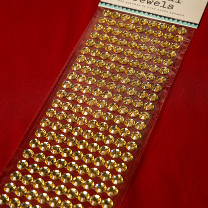 A sheet of Printable Heaven 10mm Gold Crystal Adhesive Jewels is shown on a red fabric background, ideal for embellishing crafts or adding sparkle to Christmas cards.