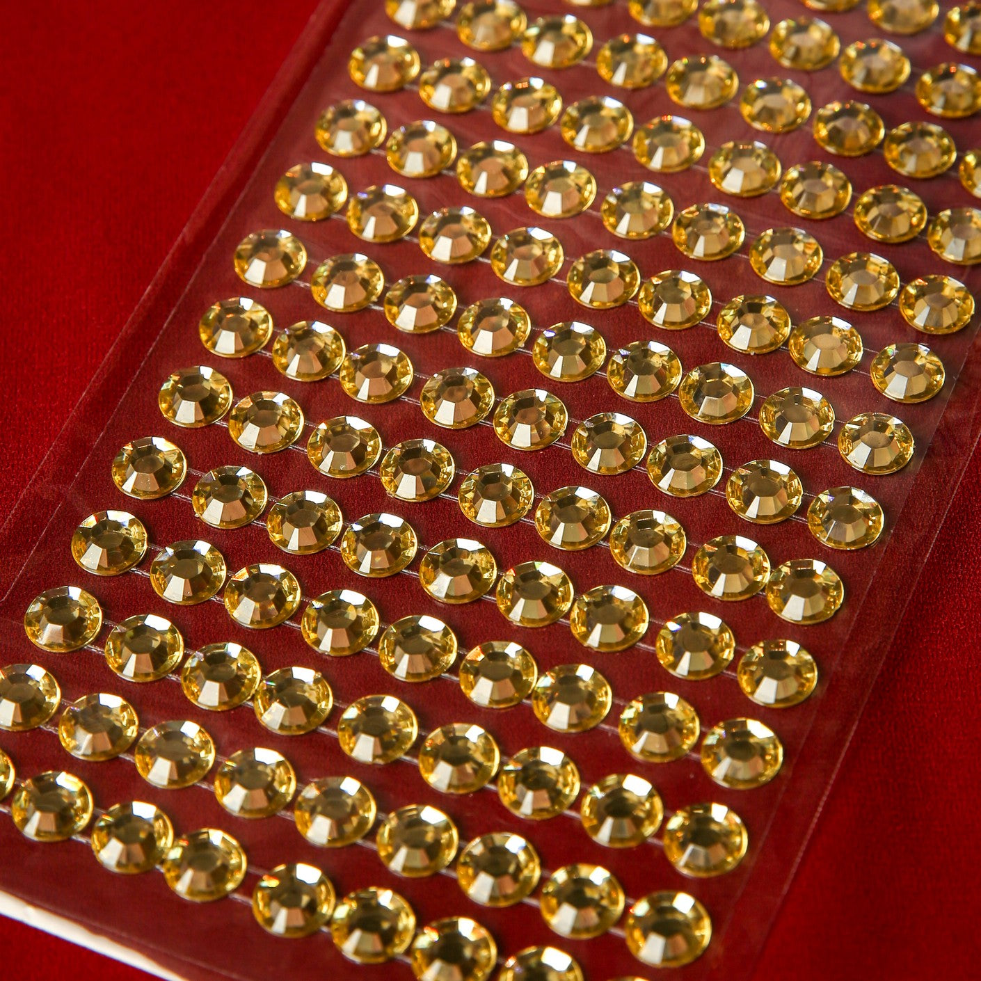 Printable Heaven's 10mm Gold Crystal Adhesive Jewels—round, faceted gems on a clear sheet—make festive embellishments for Christmas cards, shown here arranged in neat rows against a red background.
