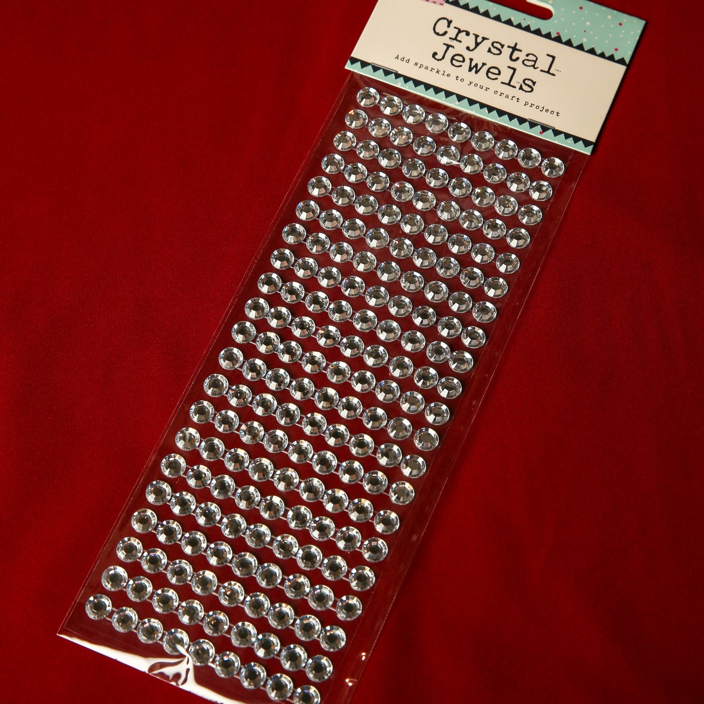 A pack of 10mm Silver Crystal Adhesive Jewels from Printable Heaven is displayed on a red surface. The clear, round gems are arranged in rows—ideal for embellishment collections or adding sparkle to Christmas cards.