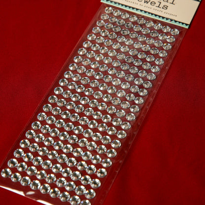 A pack of Printable Heaven 10mm Silver Crystal Adhesive Jewels is displayed on a red surface. These round, gem-like stickers are ideal embellishments for decorating Christmas cards and crafts.