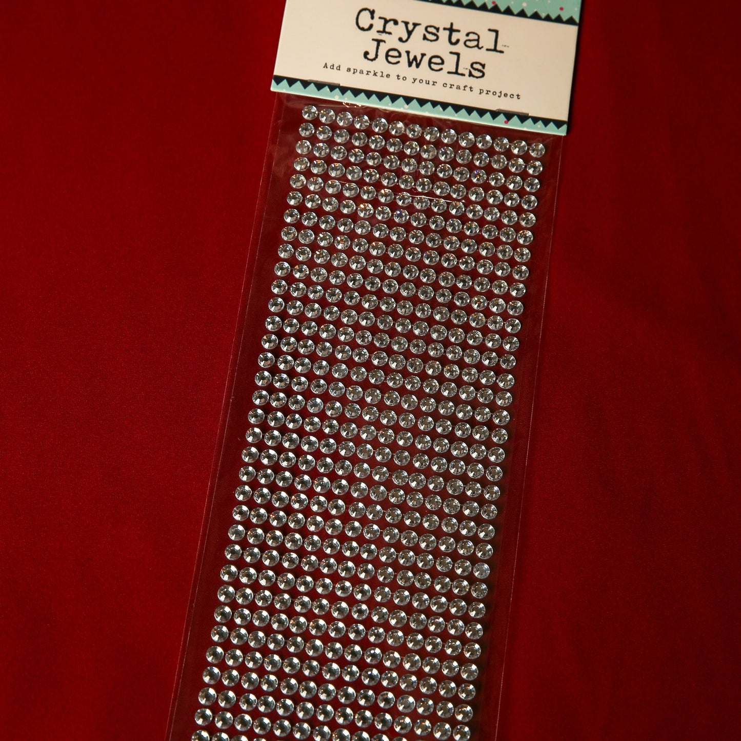 A pack of Printable Heaven 6mm Silver Crystal Adhesive Jewels arranged on a clear sheet, ideal for adding sparkle to crafts, scrapbook embellishments, or Christmas cards, shown on a red background.