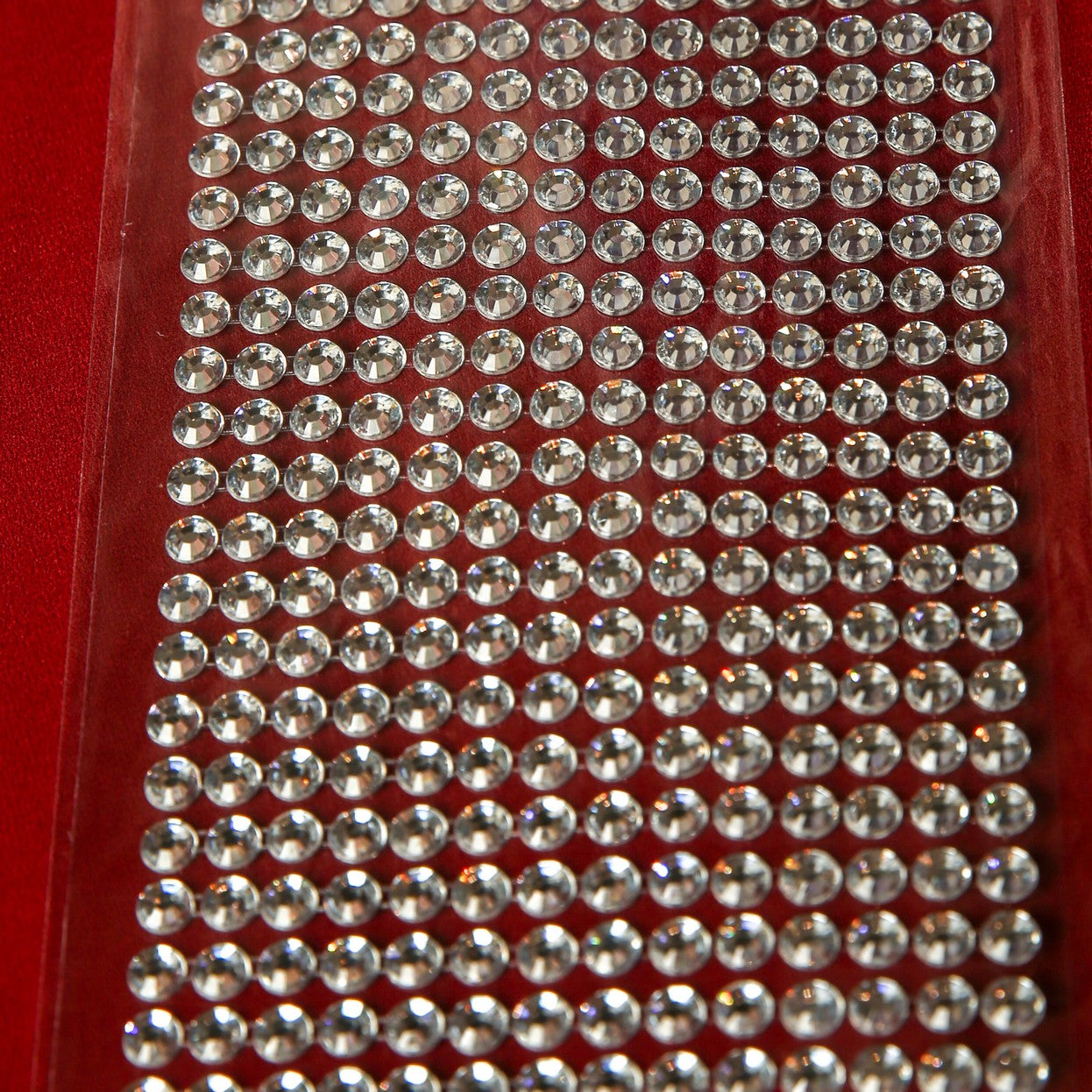 A close-up of Printable Heaven’s 6mm Silver Crystal Adhesive Jewels—small round stickers arranged in neat rows on a clear backing, ideal for scrapbooking or embellishments, shown against a red background.