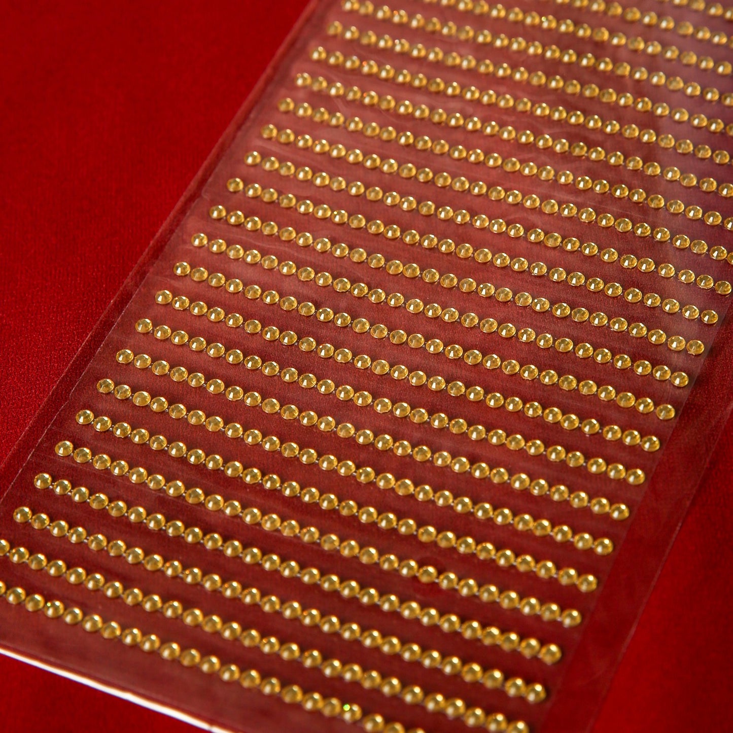 A close-up of Printable Heaven's 3mm Gold Crystal Adhesive Jewels—transparent sheets with rows of round gold gems on a red background, ideal for embellishing Christmas cards.