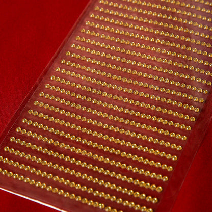 A close-up of Printable Heaven's 3mm Gold Crystal Adhesive Jewels—transparent sheets with rows of round gold gems on a red background, ideal for embellishing Christmas cards.
