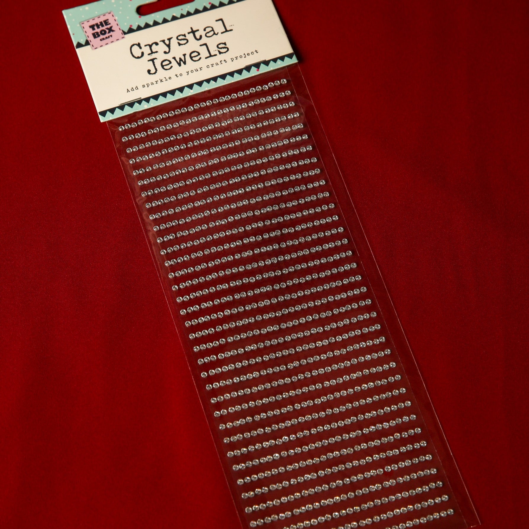 A package of Printable Heaven 3mm Silver Crystal Adhesive Jewels is displayed on a red surface, featuring rows of small, clear, round gems neatly arranged in straight lines.