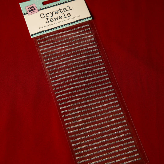 A package of Printable Heaven 3mm Silver Crystal Adhesive Jewels is displayed on a red surface, featuring rows of small, clear, round gems neatly arranged in straight lines.