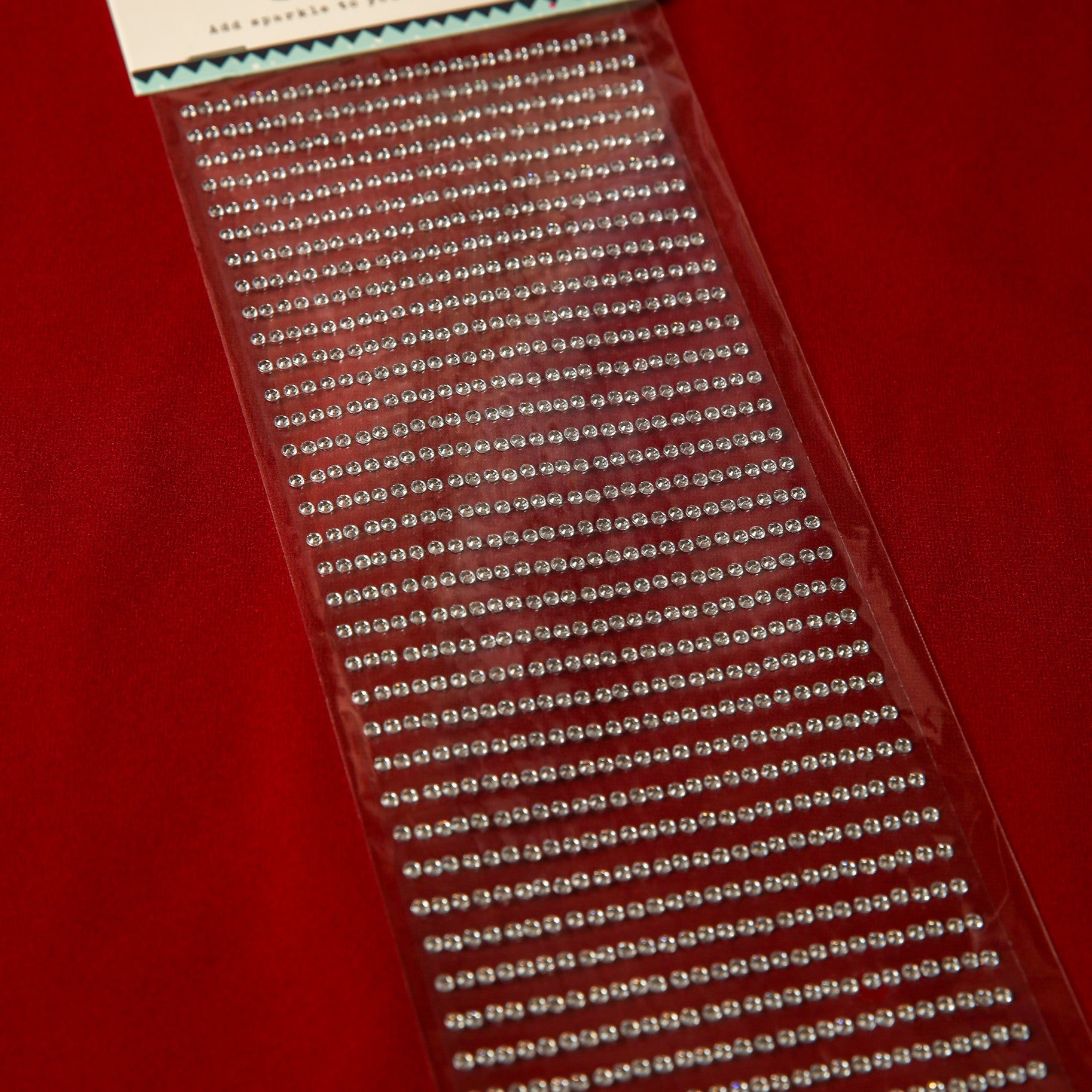 Printable Heaven’s 3mm Silver Crystal Adhesive Jewels, displayed on red fabric, come in neat rows within clear packaging—ideal for Christmas cards or scrapbook embellishments.