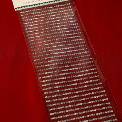 Printable Heaven’s 3mm Silver Crystal Adhesive Jewels, displayed on red fabric, come in neat rows within clear packaging—ideal for Christmas cards or scrapbook embellishments.