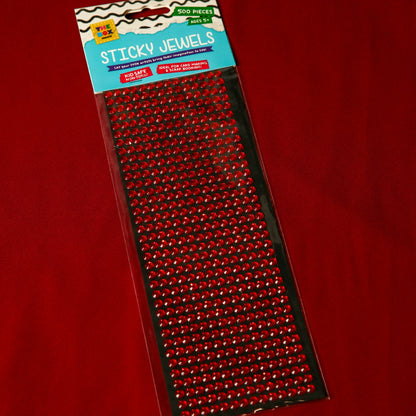 A package of 500 Printable Heaven 6mm Red Crystal Sticky Gems, ideal for crafts, lies on a red surface. The acrylic gems are lined up on black backing in clear packaging labeled Sticky Jewels.