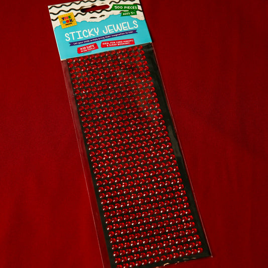 A package of 500 Printable Heaven 6mm Red Crystal Sticky Gems, ideal for crafts, lies on a red surface. The acrylic gems are lined up on black backing in clear packaging labeled Sticky Jewels.
