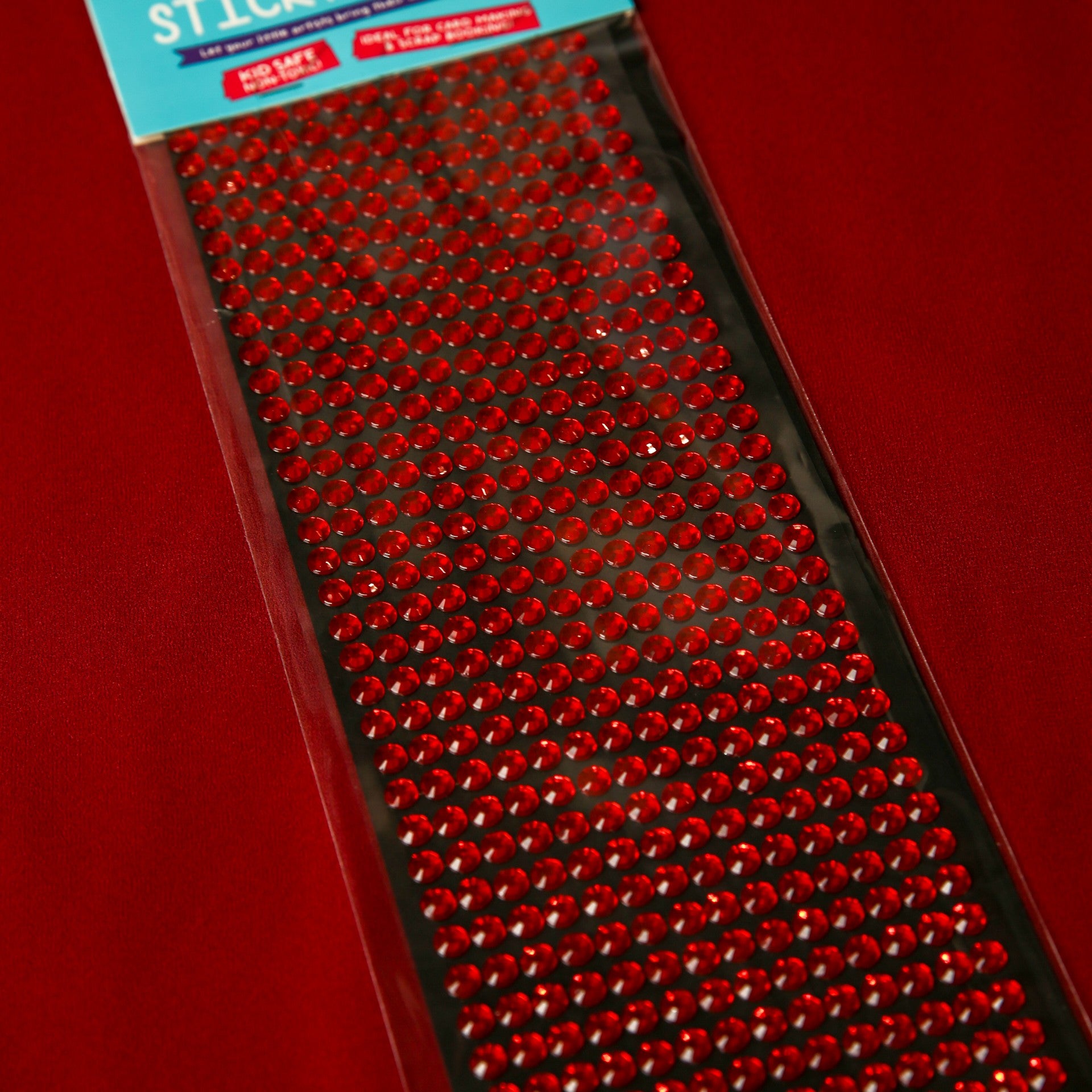 A pack of 6mm Red Crystal Sticky Gems by Printable Heaven, arranged in neat rows, perfect for craft projects, displayed on a red background.