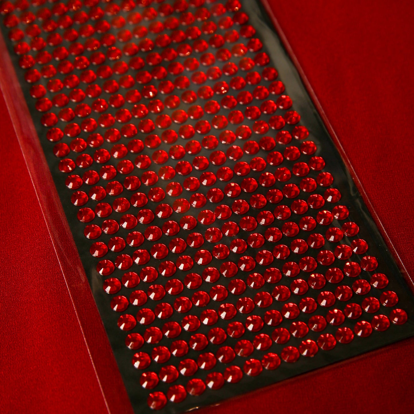 A sheet of Printable Heaven 6mm Red Crystal Sticky Gems, small round self-adhesive jewels arranged on black backing, ideal for crafts and displayed on a red surface.