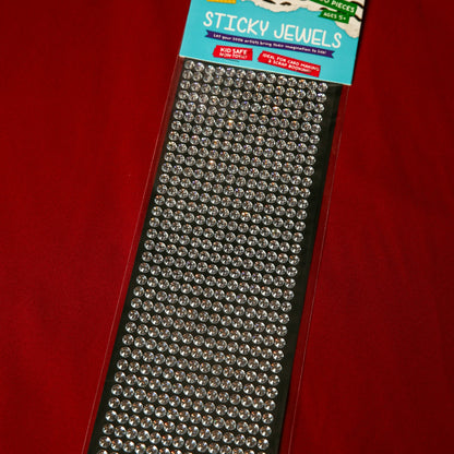 A pack of Printable Heaven 6mm Silver Crystal Sticky Gems—small, round, self-adhesive jewels on black backing—displayed on a red surface. Ideal for adding sparkle to craft projects.