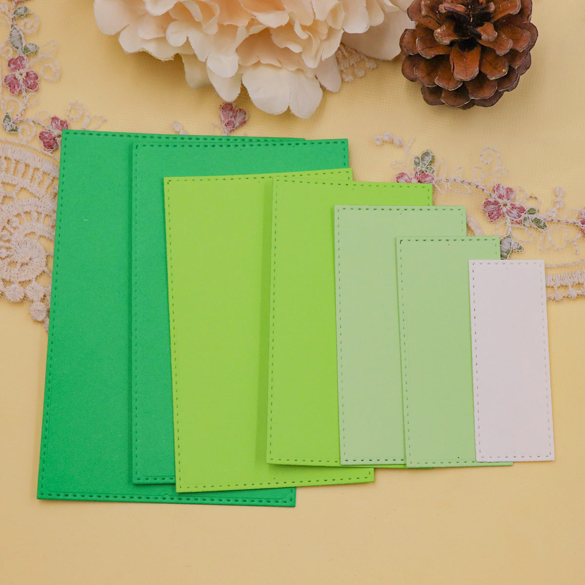 Eight rectangular shapes in shades of green and white, each showcasing an elegant stitched-edge detail created with the Printable Heaven Stitched Edge Nesting Rectangle Cutting Dies (8 PCS) (SP097), are displayed fanned out on a lace cloth surrounded by flowers and a pine cone—ideal inspiration for your next card making project.