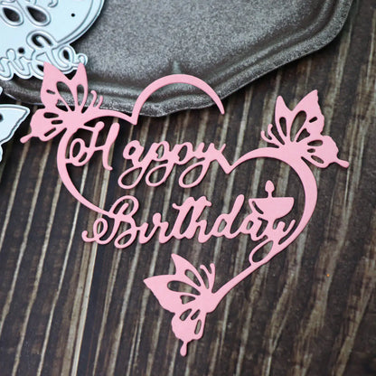 A pink, heart-shaped “Happy Birthday” cutout featuring butterfly designs—crafted with the Happy Birthday Sentiment Cutting Die - Floral (1 PC) (DJ036) by Printable Heaven—is displayed on a dark wooden surface next to a metallic tray and metal die cuts. Ideal for paper craft projects or creating handmade birthday cards.