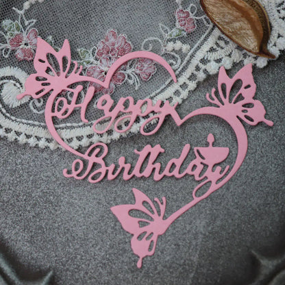 A pink “Happy Birthday Sentiment Cutting Die - Floral (1 PC) (DJ036)” from Printable Heaven, featuring delicate butterfly designs, is displayed on a gray fabric surface alongside lace and a brown button—ideal for paper craft projects and handmade birthday cards.