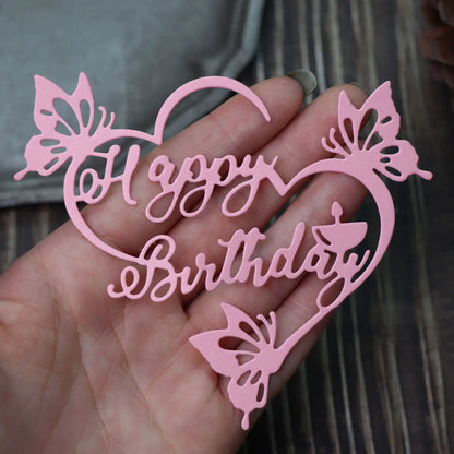 A hand holds the Printable Heaven Happy Birthday Sentiment Cutting Die - Floral (1 PC) (DJ036), a pink, heart-shaped cutout with elegant butterfly designs, displayed on a wooden surface background—an enchanting accessory ideal for papercraft projects or handmade birthday cards.