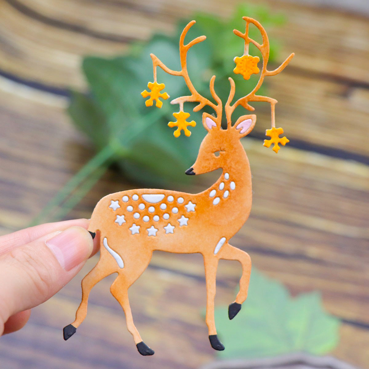 A hand holds a decorative cutout of a deer with star-shaped ornaments hanging from its antlers, crafted using the Majestic Reindeer Stag Cutting Die (1 PC) (SP013) by Printable Heaven. The blurred wood and green leaves in the background evoke the charm of woodland craft supplies.
