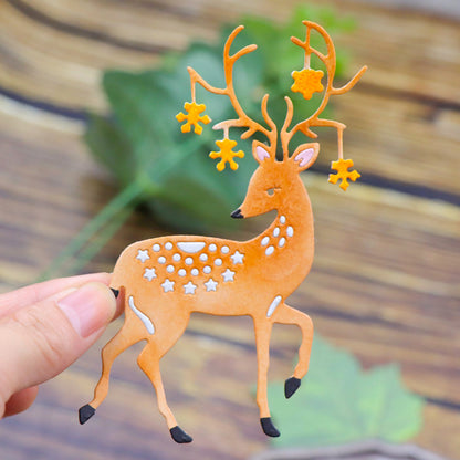 A hand holds a decorative cutout of a deer with star-shaped ornaments hanging from its antlers, crafted using the Majestic Reindeer Stag Cutting Die (1 PC) (SP013) by Printable Heaven. The blurred wood and green leaves in the background evoke the charm of woodland craft supplies.