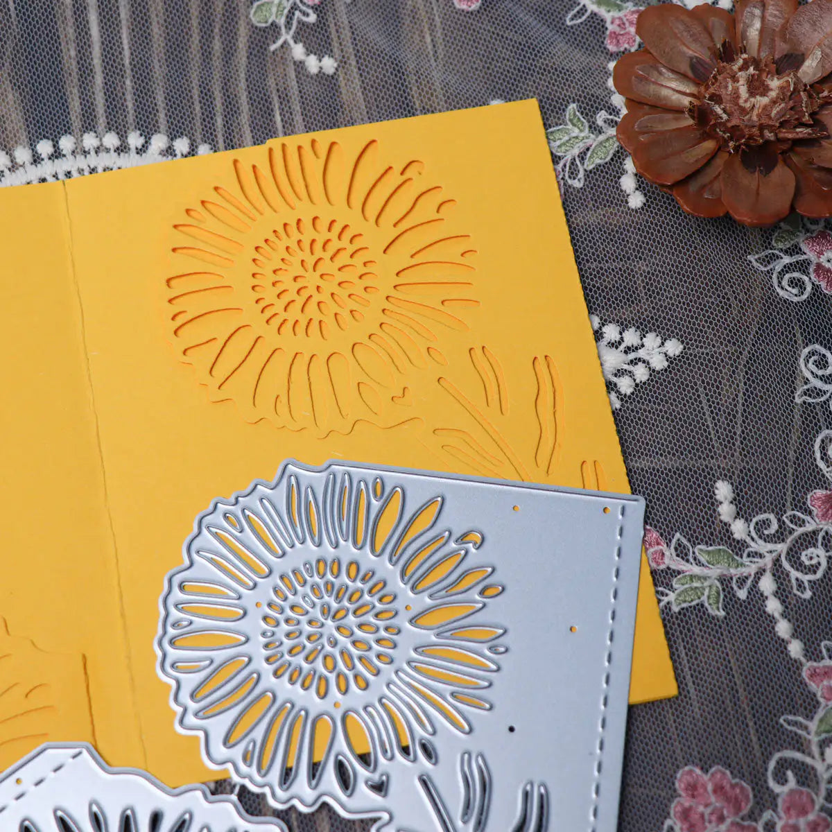 A Large Sunflower Cutting Die (2 PCS) (B049) by Printable Heaven is displayed atop a yellow card featuring a coordinating sunflower cutout, set on lace fabric with an artificial brown flower in the corner.
