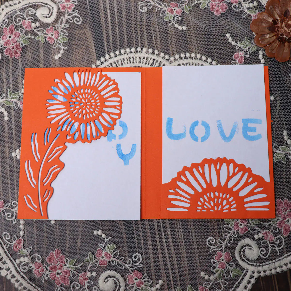 A handmade orange and white card featuring a sunflower cutout created with the Printable Heaven Large Sunflower Cutting Die (2 PCS) (B049), adorned with blue “LOVE” lettering, displayed on a lace-patterned surface decorated with pink and green florals.