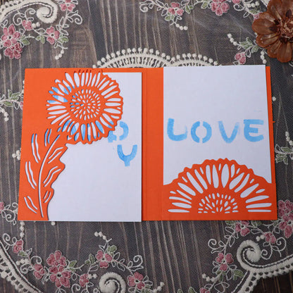 A handmade orange and white card featuring a sunflower cutout created with the Printable Heaven Large Sunflower Cutting Die (2 PCS) (B049), adorned with blue “LOVE” lettering, displayed on a lace-patterned surface decorated with pink and green florals.
