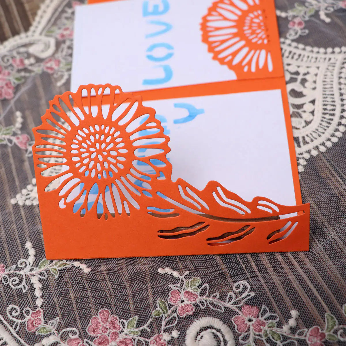 A bright orange card featuring a large sunflower design on the corner, crafted with the Printable Heaven Large Sunflower Cutting Die (2 PCS) (B049), displays “YOU LOVE” handwritten in blue inside and rests atop floral-patterned lace fabric.