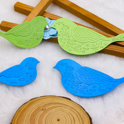 Four decorative paper birds crafted using the Printable Heaven Decorative Bird Cutting Die Set – 3D Wing Design (4 PCS) (SP126) rest on a white surface. Two green birds are placed near wooden sticks and a blue flower, while two blue birds sit by a wooden disc. Each bird showcases intricate cut-out patterns and a 3D wing design, making them ideal for papercraft projects or for highlighting your favorite cutting die techniques.