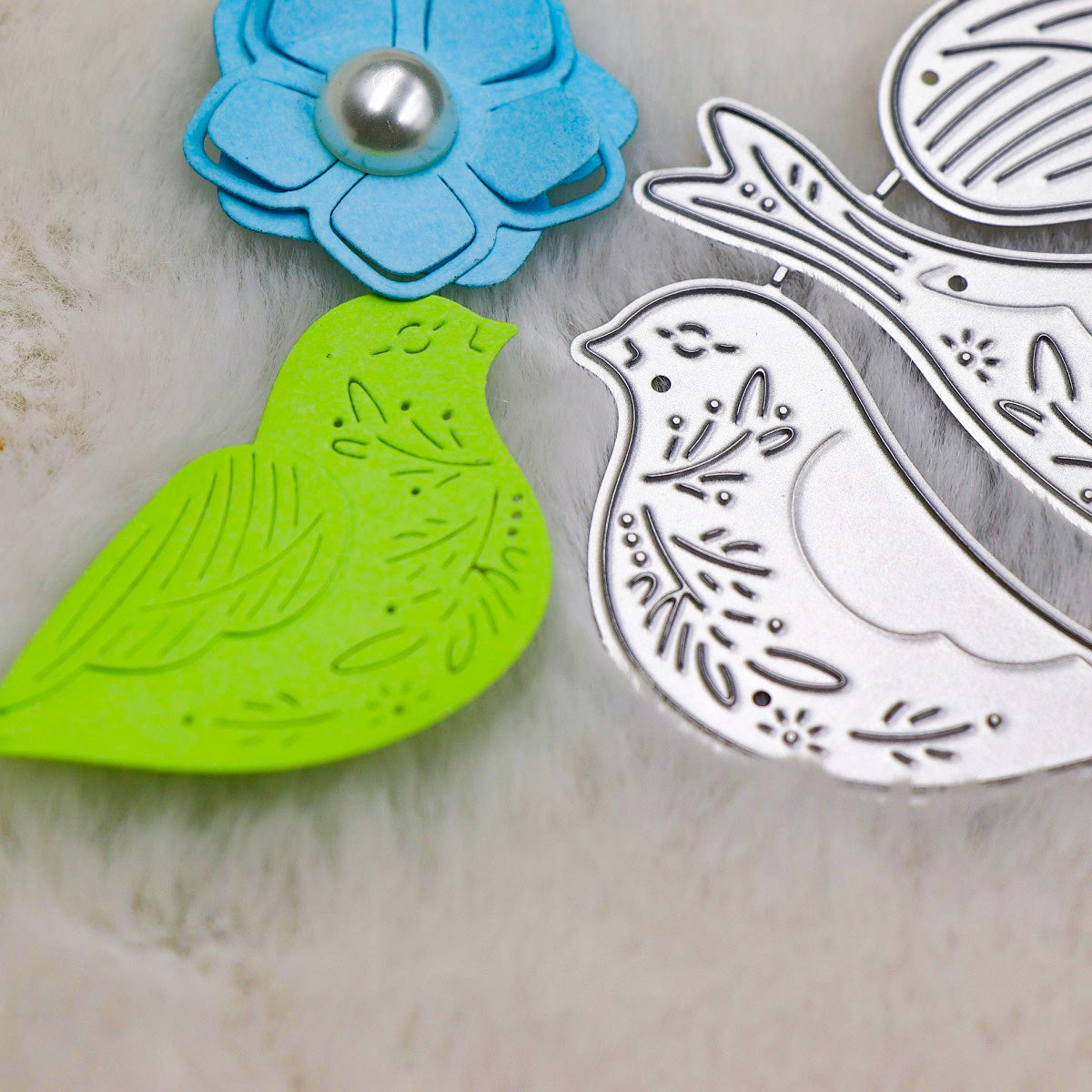 Lying on a soft white surface, the Printable Heaven Decorative Bird Cutting Die Set – 3D Wing Design (4 PCS) (SP126) includes a charming lime green paper bird with delicately layered 3D wings and two elegant silver metal dies, accompanied by a blue paper flower adorned with a pearl center—ideal accents to enrich your papercraft projects.