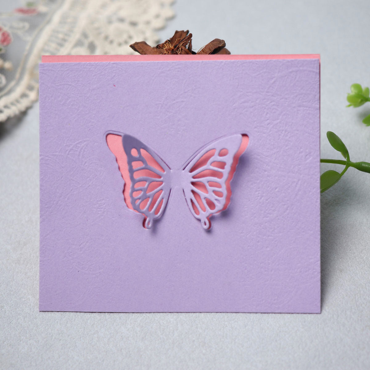 A lavender card created using the Printable Heaven Delicate Butterfly Cutting Die (2 PCS) (A188) reveals a soft pink layer beneath its intricate butterfly cutout, displayed on a light surface beside lace fabric and a green plant—an ideal source of inspiration for paper crafting enthusiasts.