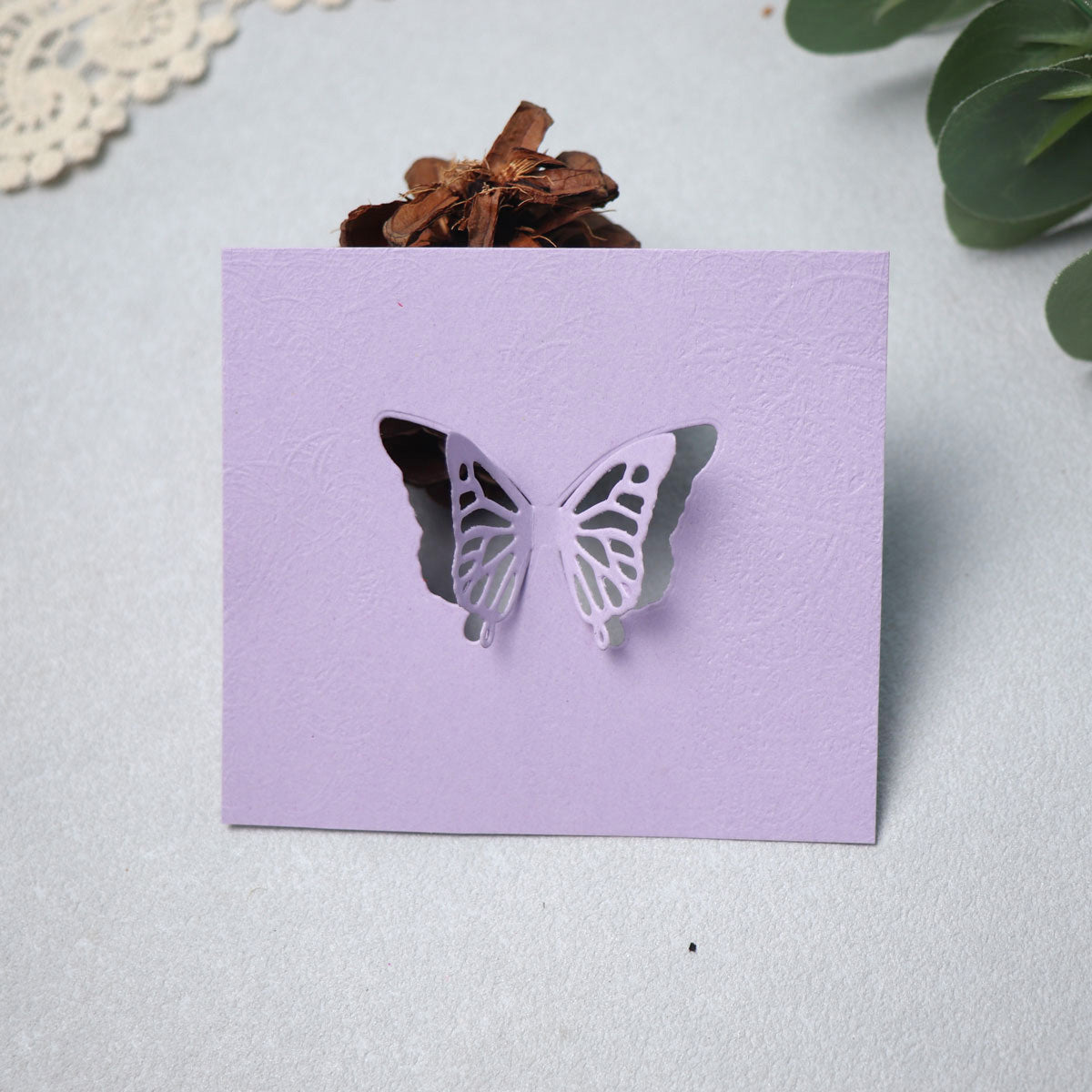 A lavender card featuring a detailed butterfly cutout, made with the Printable Heaven Delicate Butterfly Cutting Die (2 PCS) (A188), stands upright to showcase its intricate design. With green leaves and a decorative brown pinecone in the background, this scene offers perfect inspiration for your next paper crafting project.
