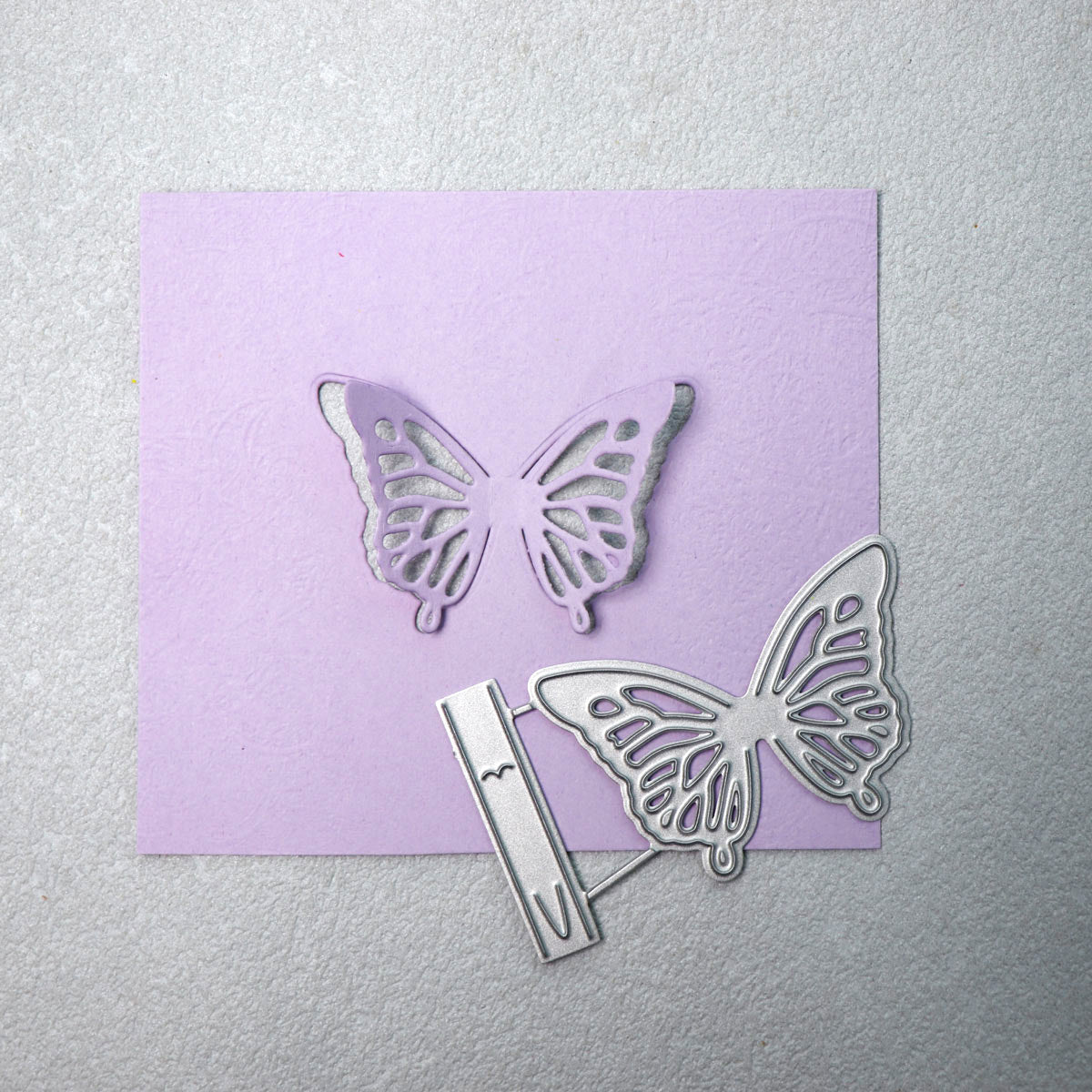 A silver Delicate Butterfly Cutting Die (2 PCS) (A188) from Printable Heaven, along with its coordinating butterfly cutout, are displayed on a purple square of textured paper atop a light gray surface—ideal for bringing an elegant touch to your next paper crafting project.