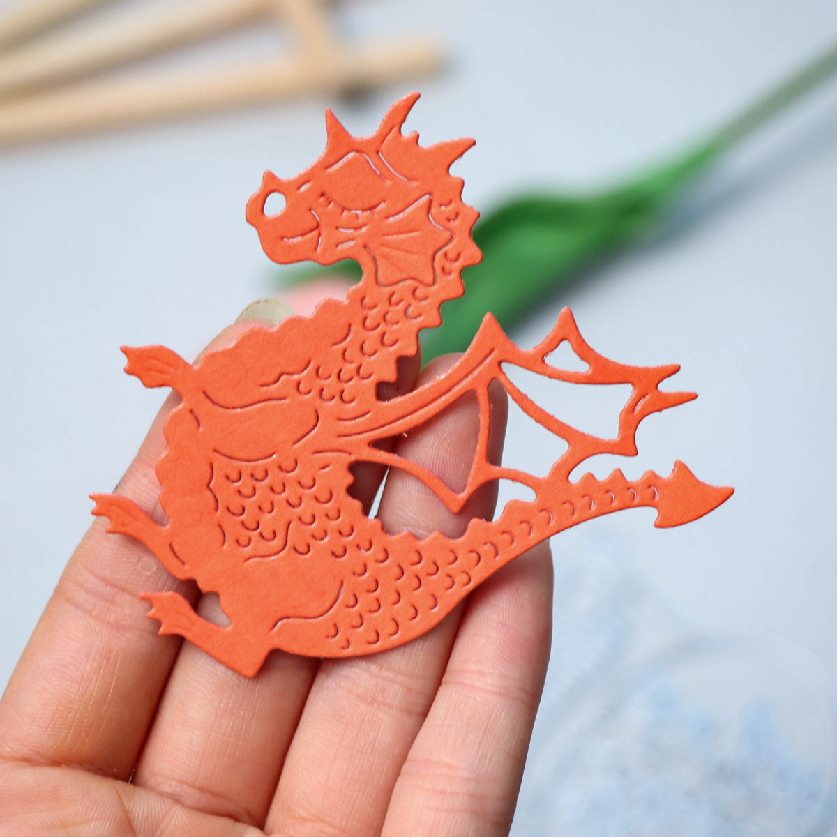 A hand holds an orange paper cutout of a dragon with a textured body and open wings, crafted using the Printable Heaven Dragon & Egg Cutting Die (3 PCS) (A111). The blurred background reveals a green leaf and wooden sticks, enhancing the whimsical atmosphere of this fantasy papercraft creation.