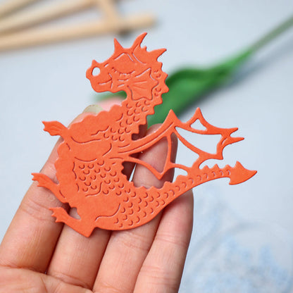 A hand holds an orange paper cutout of a dragon with a textured body and open wings, crafted using the Printable Heaven Dragon & Egg Cutting Die (3 PCS) (A111). The blurred background reveals a green leaf and wooden sticks, enhancing the whimsical atmosphere of this fantasy papercraft creation.