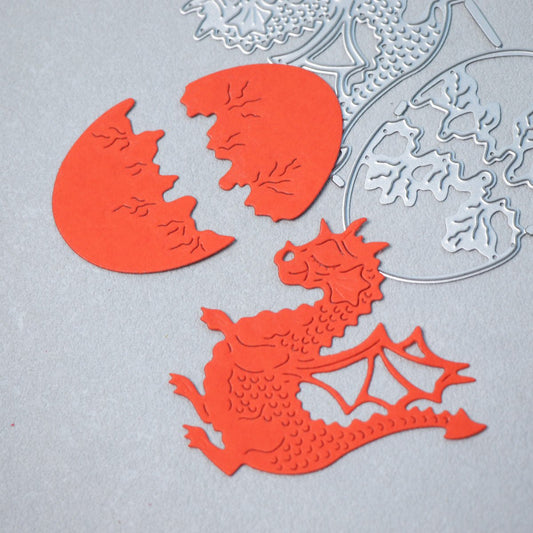Red paper cutouts of a dragon and a cracked egg lie on a gray surface, with Printable Heaven’s Dragon & Egg Cutting Die (3 PCS) set (A111)—featuring intricate fantasy designs—visible in the background.