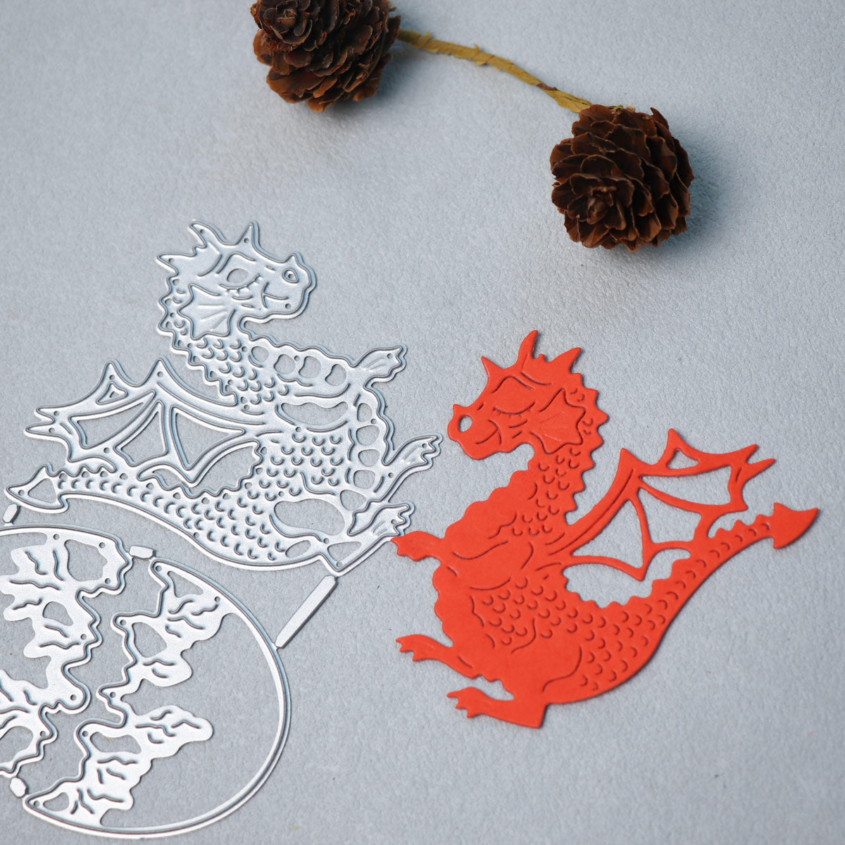 A vibrant red dragon papercut, crafted using the Printable Heaven Dragon & Egg Cutting Die (3 PCS) (A111), rests on a gray surface beside its matching die, capturing the magic of fantasy-themed papercraft. Above them, two brown pine cones on a short stem complete the enchanting scene.