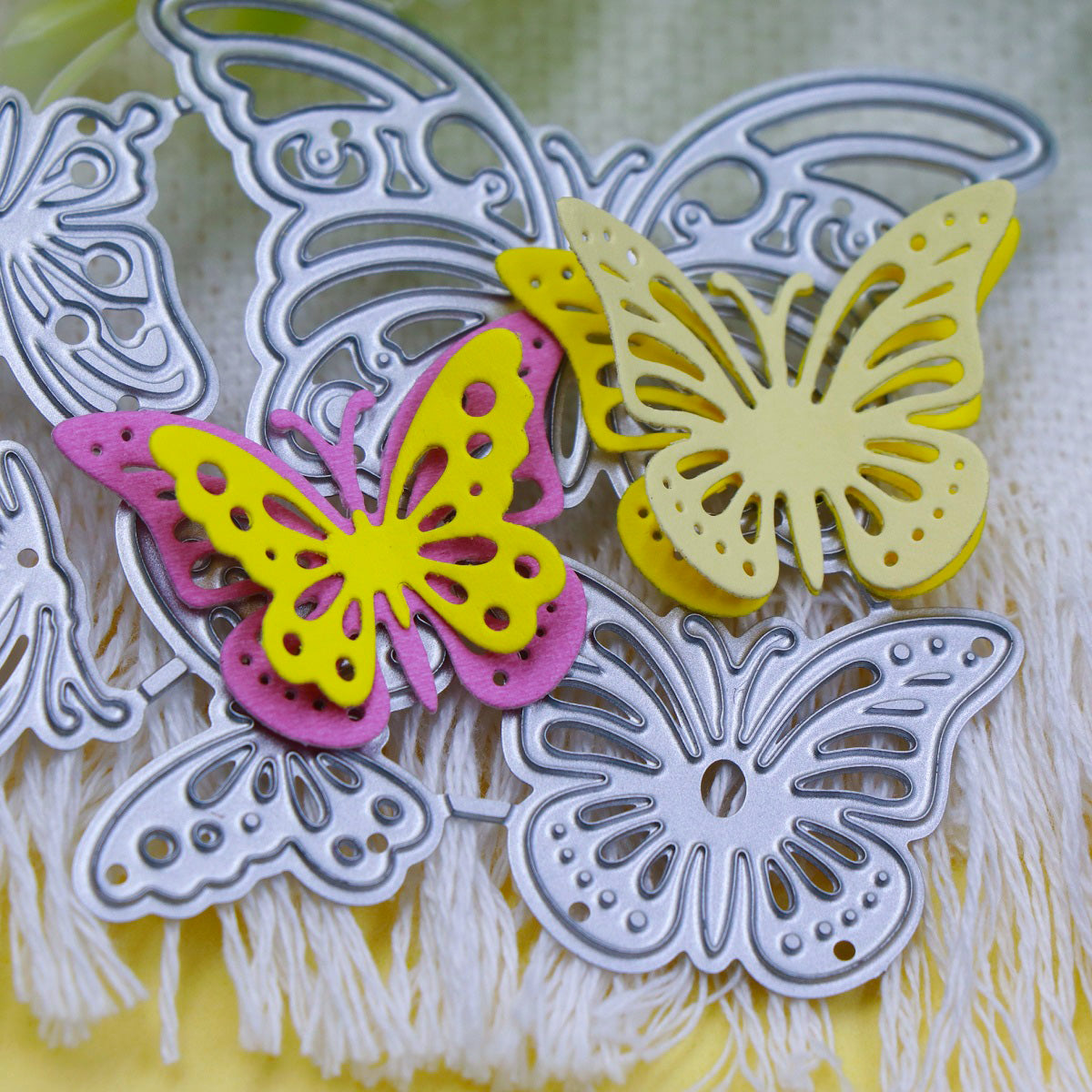 Close-up of silver metal butterfly-shaped dies from the Printable Heaven Fluttering Butterflies Cutting Die (5PCS) (A214) set, shown alongside two layered paper butterflies in yellow and pink on a textured white fabric background—an ideal addition to your cardmaking supplies.